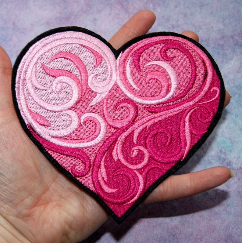 May include: A pink and white heart-shaped embroidered patch with a swirling floral design. The patch has a black border.