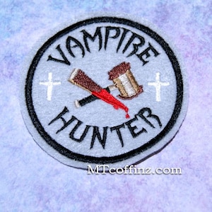May include: A white, round embroidered patch with a black border. The patch features the words "Vampire Hunter" in a black, stylized font. A crossed wooden stake and hammer with a red blood splatter are in the center of the patch. Two white crosses are on either side of the stake and hammer.