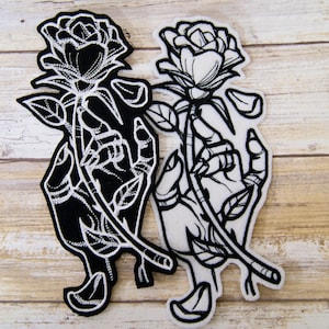 May include: Two black and white embroidered iron-on patches of a hand holding a rose. The rose is outlined in white and the hand is filled in black on one patch. The rose is filled in white and the hand is outlined in white on the other patch.