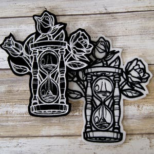 May include: Two black and white embroidered patches featuring an hourglass with roses. The hourglass is surrounded by roses with stems and leaves. The patches are on a wooden background.