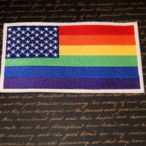 May include: A rainbow pride flag with a blue field of stars, representing the American flag, sewn onto a white background. The rainbow stripes are red, orange, yellow, green, blue, and purple.