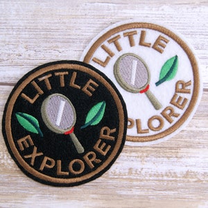 May include: Two embroidered patches with the text "LITTLE EXPLORER" in brown lettering. One patch is black with a white magnifying glass and green leaves. The other patch is white with a gray magnifying glass and green leaves.