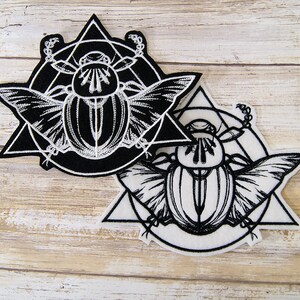 May include: Two black and white embroidered patches featuring a scarab beetle design. The beetle is surrounded by a circle and a triangle. The patches are on a white wooden background.