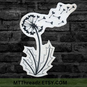 May include: Black and white embroidered design of a dandelion with seeds blowing in the wind. The design is on a white felt background.