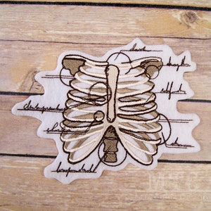 Peut inclure: Un patch brodé beige et marron représentant une cage thoracique humaine avec des étiquettes anatomiques. Les étiquettes comprennent "clavicule", "sternum", "omoplate", "humérus", "côtes" et "processus xiphoïde".