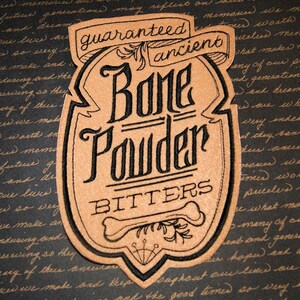 May include: A tan-colored label with black text that reads "Guaranteed Ancient Bone Powder Bitters." The label features a bone illustration and decorative flourishes. The background is a dark, textured surface with handwritten script.