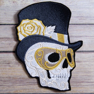 May include: Embroidered iron-on patch featuring a white skull wearing a black top hat with a gold band and a white rose. The skull has a gold and brown eye mask.