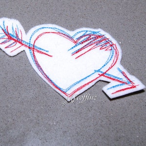 May include: White felt heart pierced by an arrow, embroidered with red and blue thread. The arrow passes through the heart, with the fletching visible on the left. The design is on a gray surface.