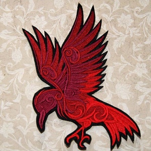 May include: Embroidered patch featuring a stylized red raven with outstretched wings. The raven has intricate detailing and a black outline. The patch is set against a cream-colored background with a floral pattern, suitable for jackets, bags, or other fabric items.