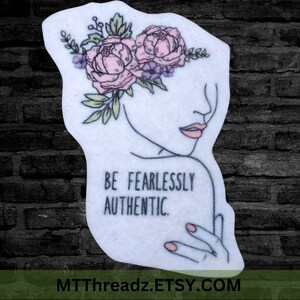 May include: A white sticker with a line drawing of a woman's face with a floral crown. The text "BE FEARLESSLY AUTHENTIC." is below the face.
