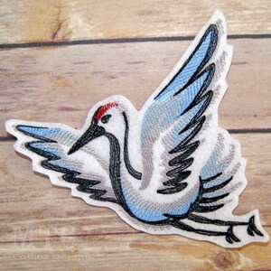 May include: Embroidered iron-on patch of a white crane with blue and gray wings, a black beak, and a red head. The crane is flying with its wings spread.