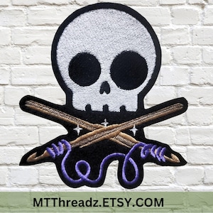 May include: A white skull with black eyes and a black mouth is crossed with two brown knitting needles. The needles are wrapped with purple yarn. The image has the text "MTTThreadz.ETSY.COM" at the bottom.