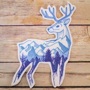 May include: A blue and white embroidered patch of a deer with a mountain range inside its silhouette.