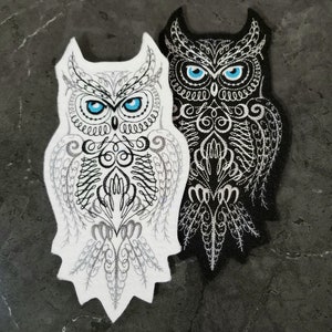 May include: Two embroidered felt owl patches, one white with black details and one black with silver details. Both owls have blue eyes and intricate designs.