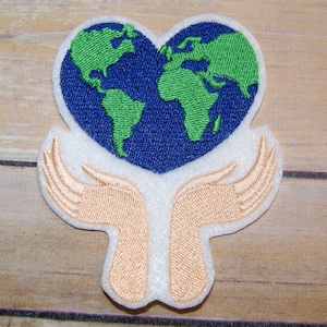 May include: Embroidered patch featuring a heart-shaped world map in shades of blue and green, held by two outstretched hands in a light peach color.
