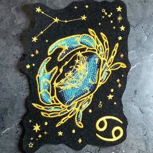 May include: Embroidered iron-on patch featuring a blue and gold crab with a constellation of stars in the background. The crab is the astrological sign for Cancer. The patch is on a black felt background.