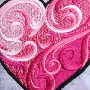 May include: A pink and white embroidered heart with a swirling pattern. The heart is outlined in black.