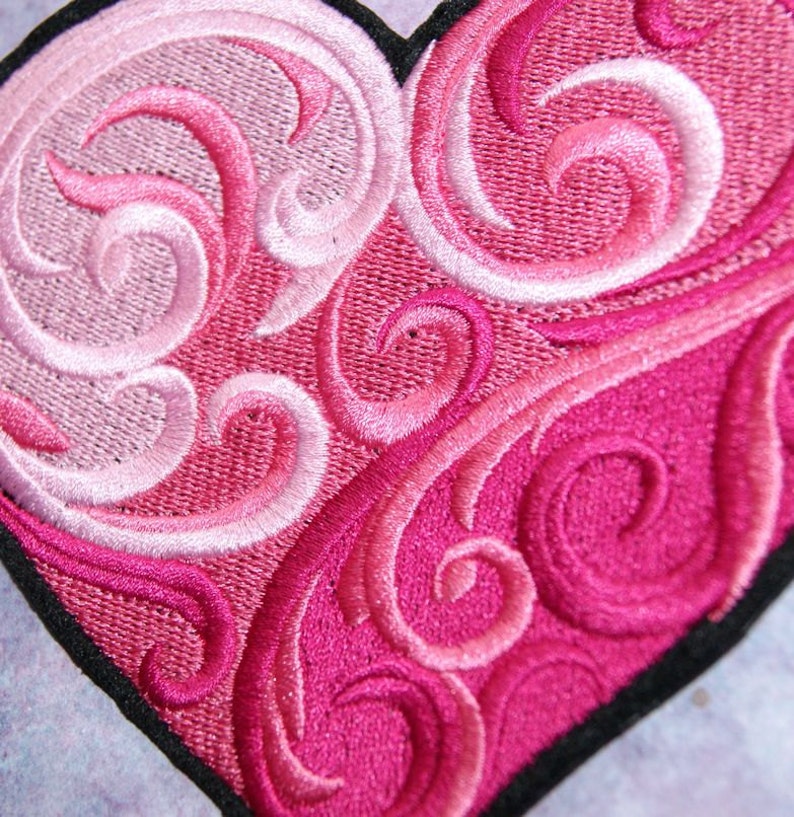 May include: A pink and white embroidered heart with a swirling pattern. The heart is outlined in black.