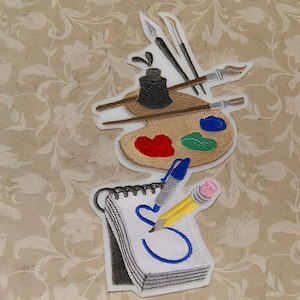May include: Embroidered patch featuring an artist's palette with paintbrushes, a paint jar, and a notepad with a pencil and pen. The palette has red, green, and blue paint on it.
