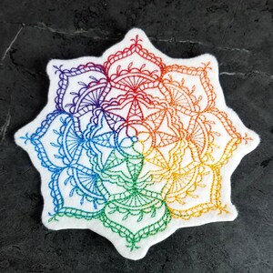 May include: A white fabric circle with a rainbow mandala design embroidered in colorful thread. The mandala features intricate patterns and details.