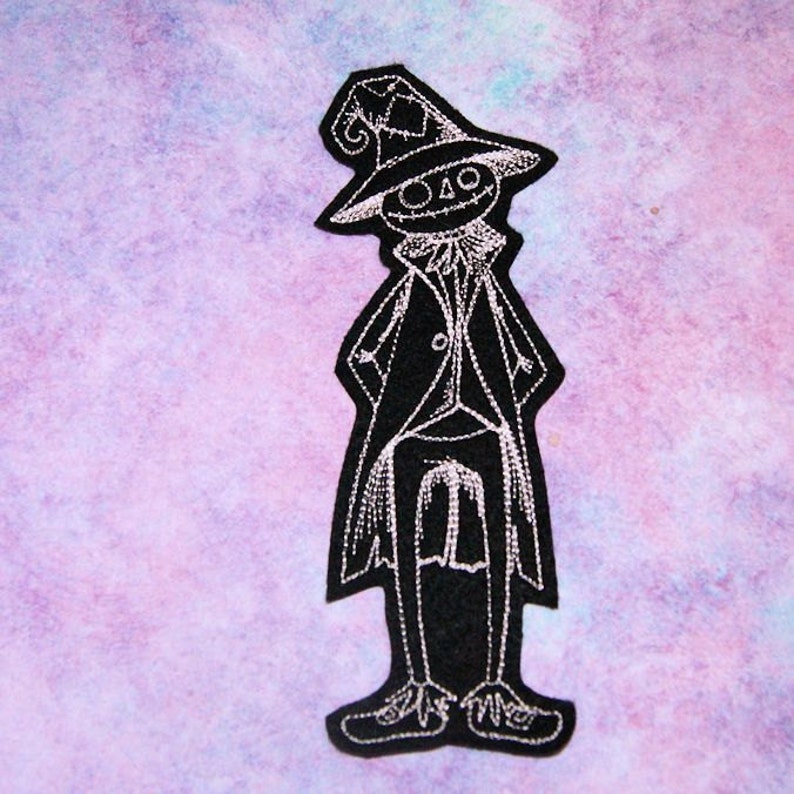 May include: Black felt embroidered patch of a skeleton wearing a tall hat and a long coat. The skeleton has a stitched smile and is standing with its hands on its hips.