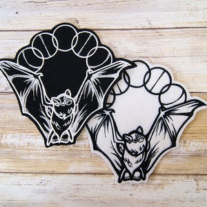 May include: Two black and white embroidered patches of a bat with wings spread wide. The bat is surrounded by a circle of five smaller circles. One patch is on a black background and the other is on a white background.