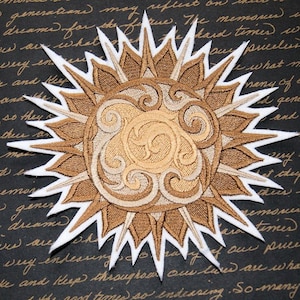 May include: A brown and white embroidered patch with a sun design. The sun has a swirling pattern in the center and is surrounded by a ring of white points.
