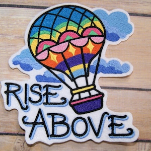 May include: Embroidered iron-on patch featuring a colourful hot air balloon and clouds. The text "RISE ABOVE" is embroidered in blue below the balloon.