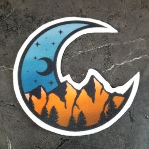 May include: A circular sticker featuring a crescent moon and mountain range design. The top half of the moon is a gradient of blue and dark blue with stars. The bottom half shows orange mountains and dark green trees.