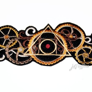 May include: A black and gold embroidered patch featuring a triangle design with a red eye at its centre. The triangle is surrounded by gears and swirls.