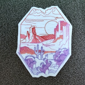 May include: Embroidered patch with a desert landscape scene. The patch features a red and pink sunset, mountains, cacti, and a large sun in the sky. The patch is outlined in purple thread.
