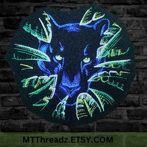 May include: Black panther head embroidered on a dark fabric patch with green and blue accents. The panther's eyes are green and the background is a dark green and blue swirl.