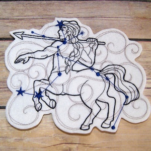 May include: A white felt patch with an embroidered outline of the constellation Sagittarius. The constellation is depicted as a centaur holding a bow and arrow. The stars are blue and the lines are black.