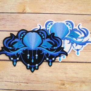 May include: Two embroidered patches featuring a blue seashell with blue and teal floral designs. The patches are on a wooden surface.