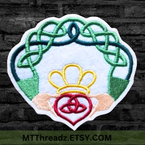 May include: An embroidered patch with a green Celtic knot design, a red heart with a trinity knot inside, and a yellow crown above. The heart is held by two hands, and the design is surrounded by a white border.