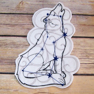 May include: A white felt patch with an embroidered outline of a wolf howling at the moon. The wolf is made up of a constellation of stars with blue stitching. The background is a swirl pattern.