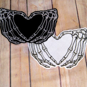 Peut inclure: Deux patchs brodés en forme de mains formant un cœur. Un patch est noir avec des coutures blanches, l'autre est blanc avec des coutures noires.