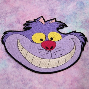May include: Purple embroidered patch of the Cheshire Cat from Alice in Wonderland, with a wide grin and yellow eyes.