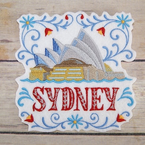 May include: Embroidered patch featuring the Sydney Opera House in white, gray, brown, and yellow with blue water. The word "SYDNEY" is embroidered in red with silver accents and surrounded by blue and red floral designs.