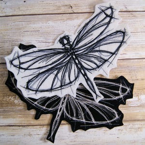 May include: Black and white embroidered dragonfly design on a white felt background. The dragonfly is outlined in black thread and has a detailed wing pattern.
