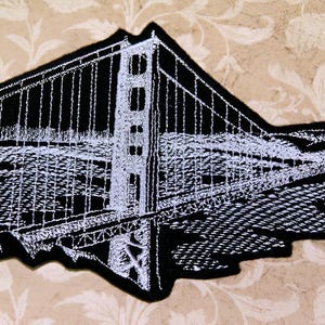 May include: Black and white embroidered patch of the Golden Gate Bridge. The patch is made of felt and has a white outline of the bridge.