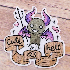 May include: Embroidered patch featuring a cute cartoon devil with pink and purple wings, holding a trident. The devil is surrounded by hearts and stars. The text "cute as hell" is written on a banner.