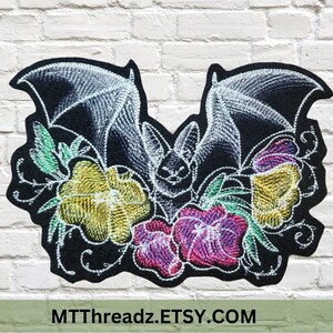 May include: Embroidered patch with a bat design, surrounded by flowers in shades of yellow, pink, and purple. The bat is white, set against a black background. The patch is displayed against a white brick wall.