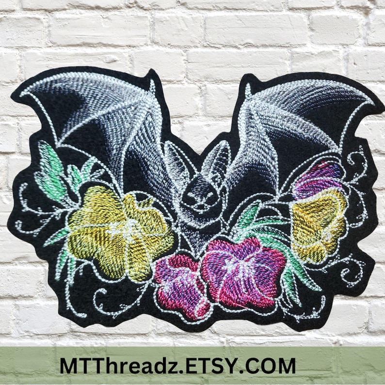 May include: Embroidered patch with a bat design, surrounded by flowers in shades of yellow, pink, and purple. The bat is white, set against a black background. The patch is displayed against a white brick wall.