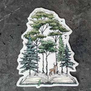 May include: An embroidered patch featuring a forest scene with trees and a deer standing on an open book. The patch has a white border and is primarily green and brown, with black detailing. The background is a dark gray, textured surface.