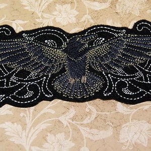 Peut inclure: Patch en feutre noir avec un motif brodé détaillé d'un corbeau aux ailes déployées. Le corbeau est entouré de motifs floraux complexes.