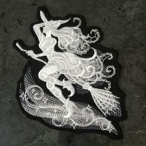 May include: White embroidered patch of a witch flying on a broomstick with a swirling design around her. The patch is on a black background.