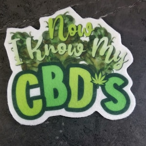 Puede incluir: Una pegatina blanca con letras verdes y negras que dicen "I Know My CBD's". Las letras están rodeadas de una imagen de hojas de marihuana de estilo acuarela verde.