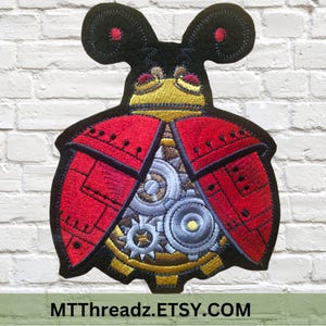 May include: An embroidered iron-on patch depicting a stylised ladybug with gears and cogs inside its body. The ladybug is red and black with black antennae and red dots on the ends. The text "MTTThreadz.ETSY.COM" is at the bottom of the image.