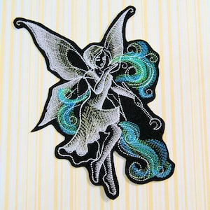 May include: White embroidered fairy with green and blue swirling designs on a black fabric background.